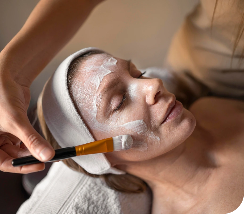Aesthetician applying a white facial mask with a brush to a patient's face during a skincare treatment